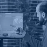 Man in a brick-walled office pointing at bar and pie charts on a large monitor, shown in a navy blue tint with the Kanerika logo