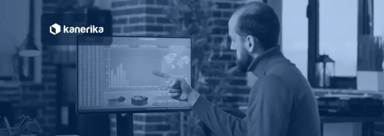 Man in a brick-walled office pointing at bar and pie charts on a large monitor, shown in a navy blue tint with the Kanerika logo