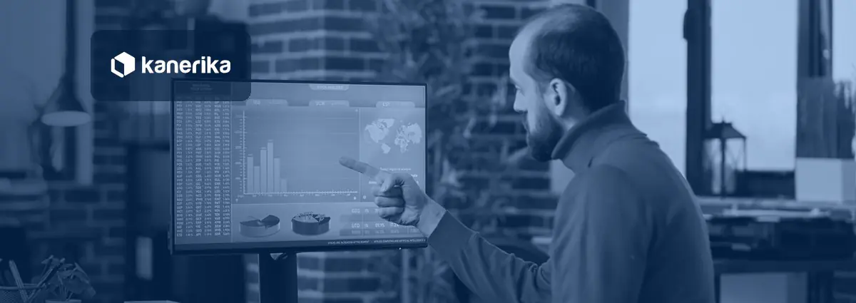 Man in a brick-walled office pointing at bar and pie charts on a large monitor, shown in a navy blue tint with the Kanerika logo