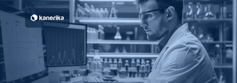Scientist in a lab coat and safety glasses reviewing data charts on computer screens in a pharmaceutical laboratory