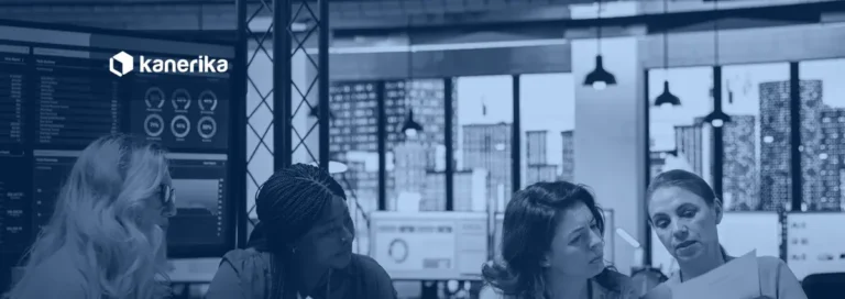 Four women gathered around a table in an office, reviewing data and reports with dashboards on a screen behind them, a team working on data governance