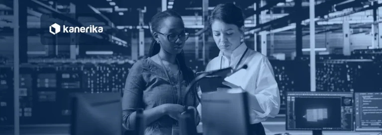 Two data engineers reviewing a tablet beside workstations in a data center, with rows of servers behind them