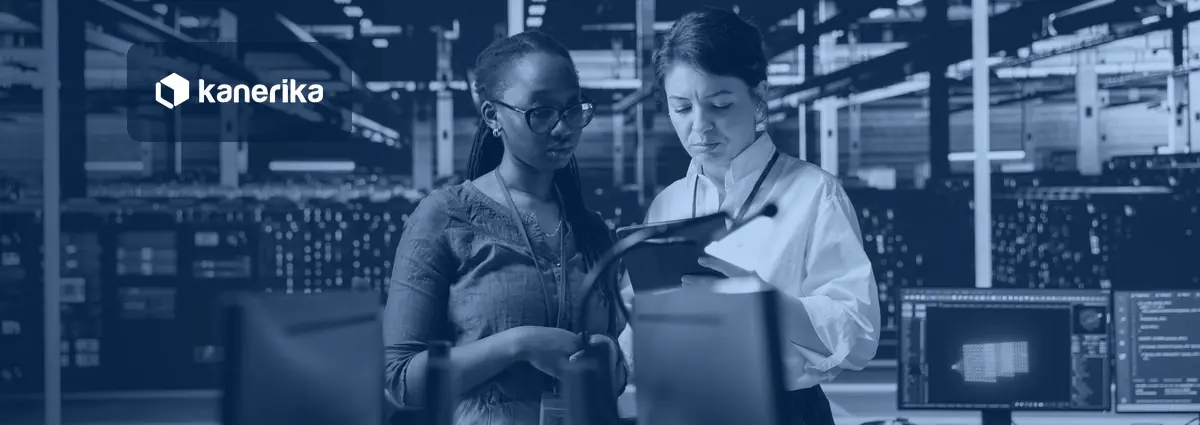 Two data engineers reviewing a tablet beside workstations in a data center, with rows of servers behind them