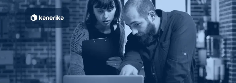 Two colleagues in a brick-walled office, one holding a clipboard, looking closely at a laptop while reviewing business paperwork